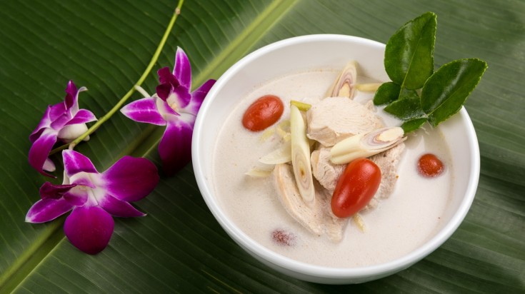 A beautiful display of Tom Kha Kai in a bowl next to purple flowers set on a banana leaf. 