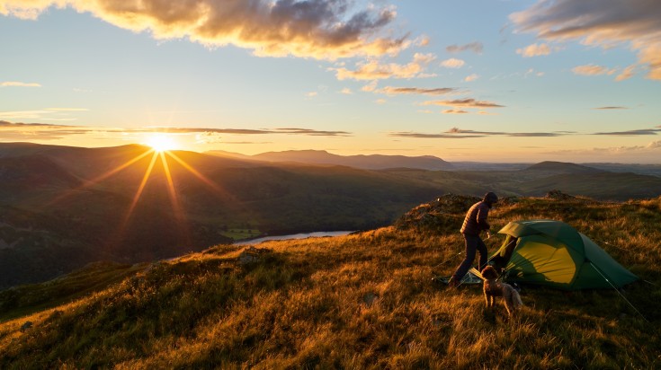 A dog looking at a man setting up a camp in the Lake District.