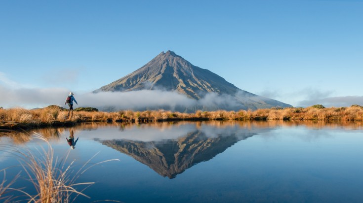 A woman hiking in front of Mount Taranaki in New Zealand in summer.