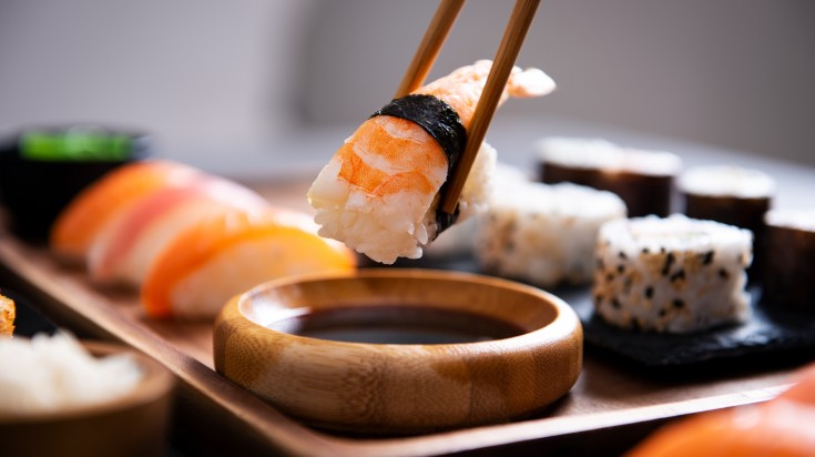 A closeup of bamboo chopsticks dipping nigiri shrimp in soy sauce.