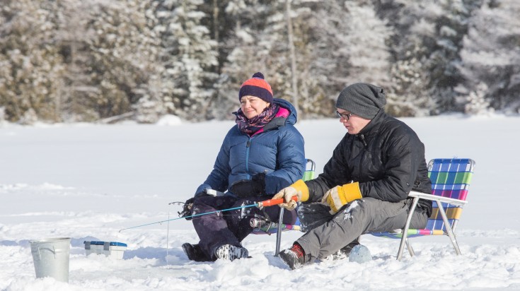 A couple sitting on frozen, snowy lake with fishing rods in hands