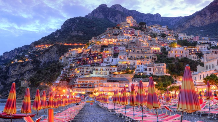 Rows of sunbed with closed colorful umbrellas and the view of illuminated houses and mountains in the background in the Amalfi Coast. 