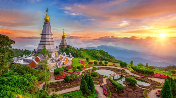 A view of ancient temples surrounded by gardens and trees with the orange sun in the distance in Chiang Mai. 