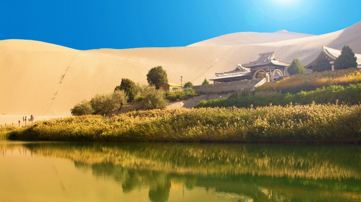 A traditional building surrounded by sandy desert, grasses and a lake