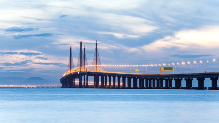 See Penang Bridge illuminated at dawn while spending one week in Malaysia.