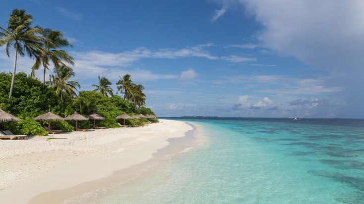 A tropical beach with white sand, clear turquoise waters, and thatched umbrellas under palm trees, set against a partly cloudy sky in the Maldives in October.