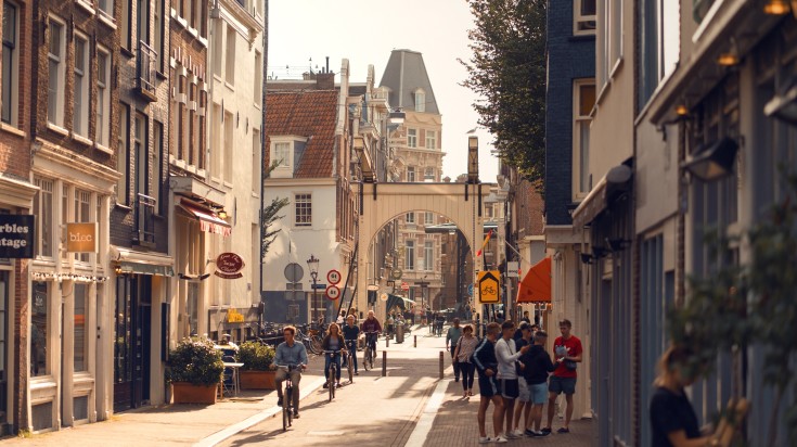 A street in Amsterdam with people walking and cycling on a bright day.