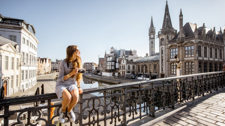 Young woman tourist sitting on the bridge of the old town of Ghent, one of the top places to visit in Belgium, on a sunny day.