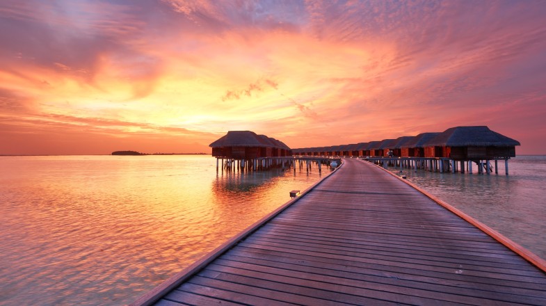 A wooden boardwalk leads to overwater bungalows at sunset in the Maldives in October, with vibrant pink and orange hues in the sky reflecting on the calm sea.