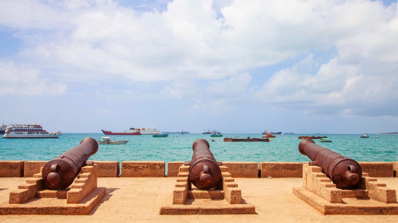 Three antique cannons lined up next to each other and positioned towards turquoise sea with ships and boats—one of the best things to do in Zanzibar.