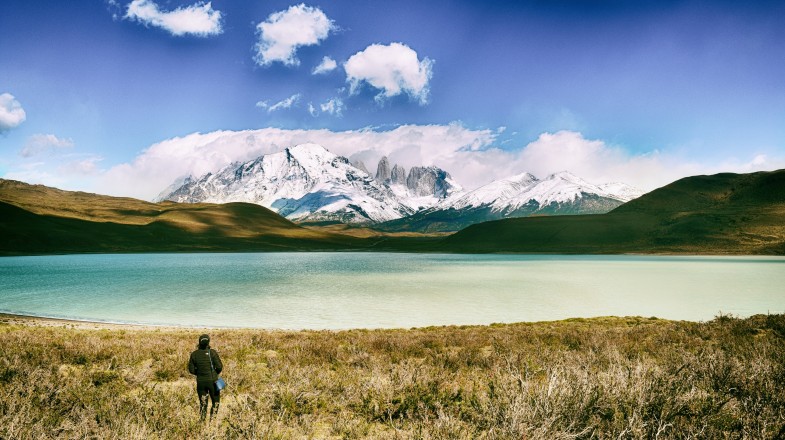 A trekker stands on a lakeshore and admires view of snow-covered mountain in Chile
