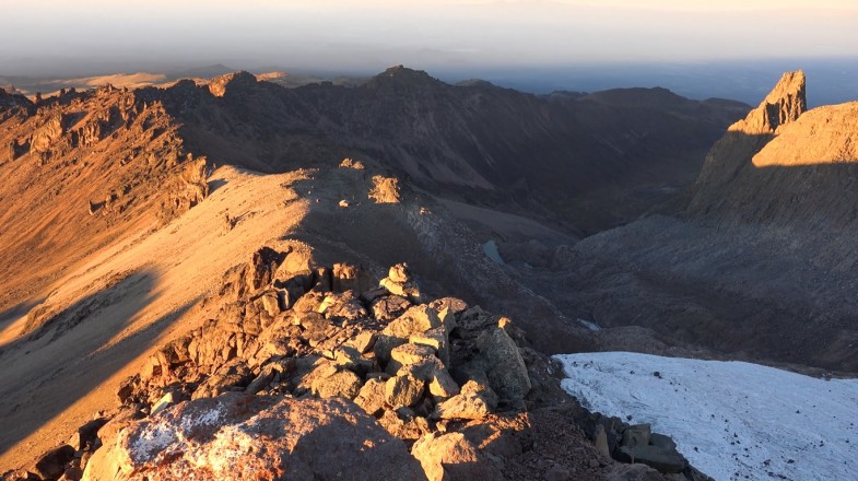 A dry, stony, barren summit of a mountain range with some snow visible on the side.